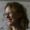 A smiling woman with wavy red hair and glasses stands in front of a whiteboard covered in mathematical equations and diagrams, wearing a black top.