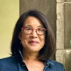 A woman with shoulder-length dark hair and red glasses stands beside a stone wall, wearing a denim jacket over a striped shirt and smiling gently at the camera.