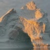 A massive iceberg rises from still, dark water, its icy surface bathed in warm golden light from a low sun under a cloudy sky.