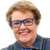 A smiling older woman with short light brown hair and bright blue glasses stands with arms crossed, wearing a denim jacket against a plain white background.