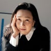 A woman with long dark hair and hoop earrings rests her face against one hand while leaning on a table, wearing a black sweater with a white collar in a softly lit indoor setting.