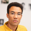 A young man, who is wearing glasses and a buttoned-down shirt, stands in front of a wall covered in photographs.