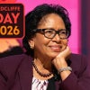 A woman wearing glasses and a maroon blazer smiles while resting her chin on her hand during a stage event, with a “Radcliffe Day 2026” graphic in the corner and a pink-lit background.
