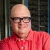 A smiling man wearing glasses leans against a concrete column.