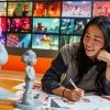 A woman sits at a table sketching colorful animated characters, surrounded by artwork and 3D character models, with a vibrant storyboard wall in the background.