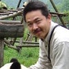 A smiling man is holding a young, furry panda bear within an outdoor animal enclosure. 