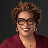 A smiling woman with short curly hair wears oversized glasses, gold earrings, and a burgundy sweater against a dark background.