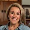 A smiling woman with short blonde hair wears a light denim shirt and pearl necklace, standing indoors with wooden bookshelves and framed photos in the background.