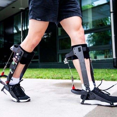 Person walking outdoors wearing robotic exoskeleton braces on both legs.