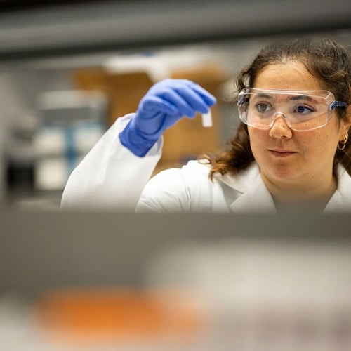Olivia Hogan-Lopez in a lab coat and safety goggles examines a small sample vial