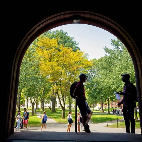 Two students in silhouette talk under an archway overlooking a bright, tree-filled Harvard Yard.